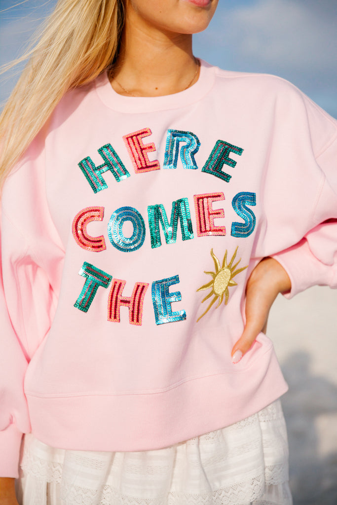 Pink cropped pullover with Here Comes The in alternating rainbow sequin letters and a sun patch