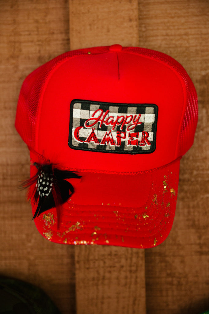 Red trucker hat with "happy camper" patch, black feathers, and glitter spray. 