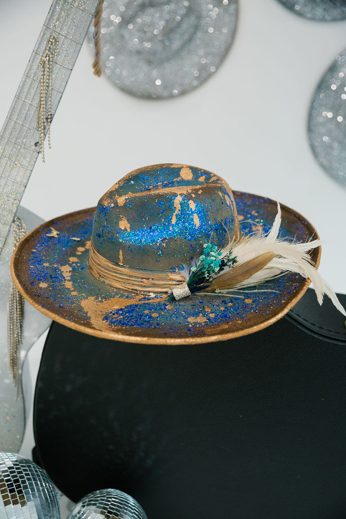 Blue rancher hat with gold accents, blue glitter spray, and feathers
