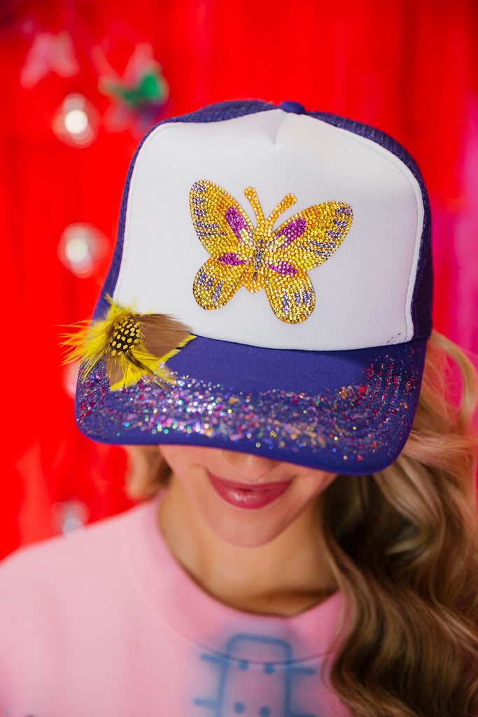 Purple and white trucker hat with a gold sequin butterfly, yellow feathers, and multi colored glitter spray