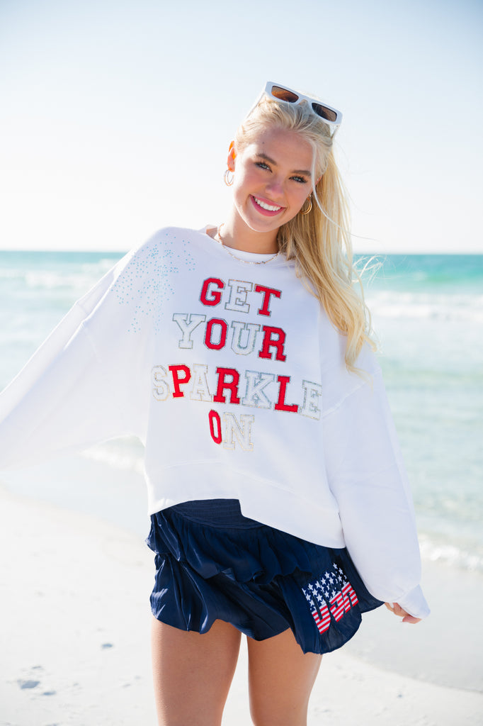 White cropped pullover with "Get Your Sparkle On" in red and white letters with blue rhinestone spray