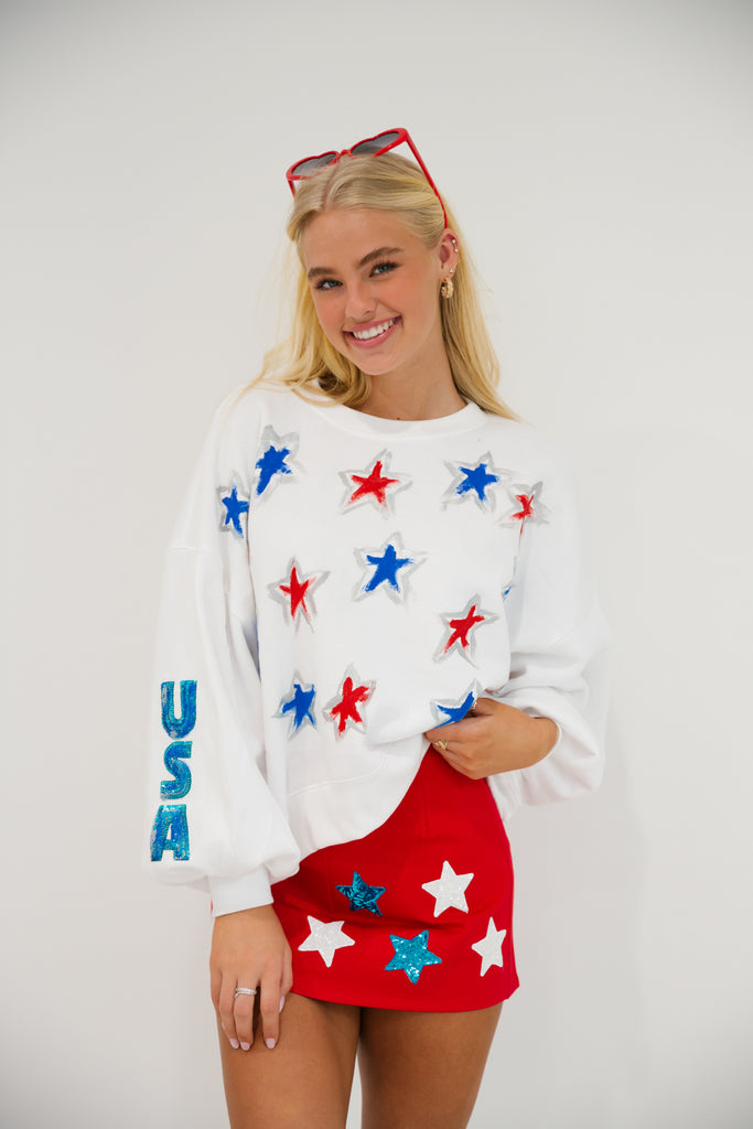 White cropped pullover with red and blue painted stars and blue sequin "USA" letters on the sleeve