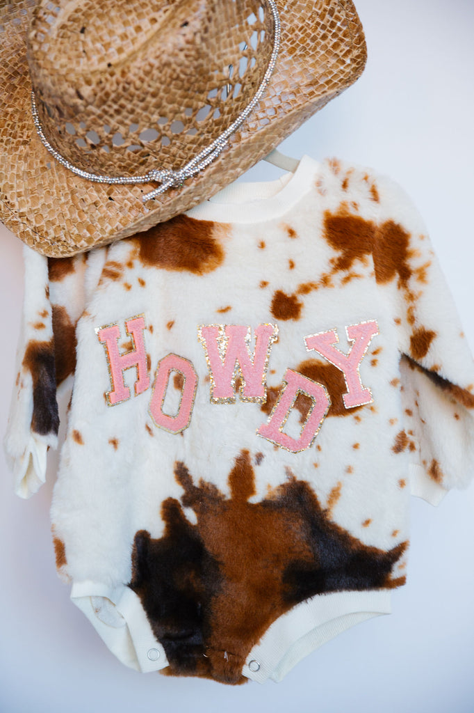 Cow print sherpa onesie with "howdy" in mini pink terry letters. 